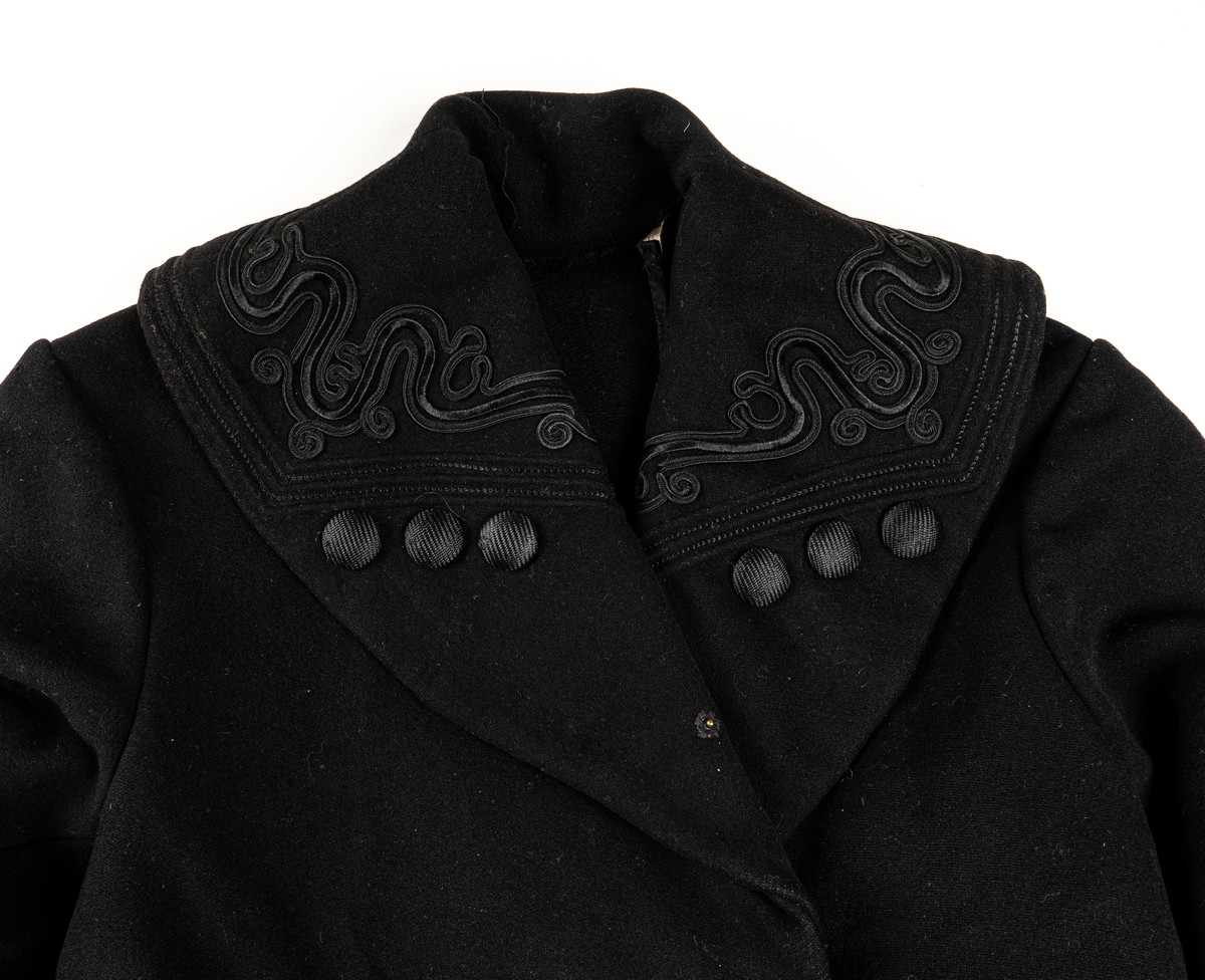 Svart kappa. Sjalkrage med snörbroderier och klädda knappar. Större oklädda knappar framtill.