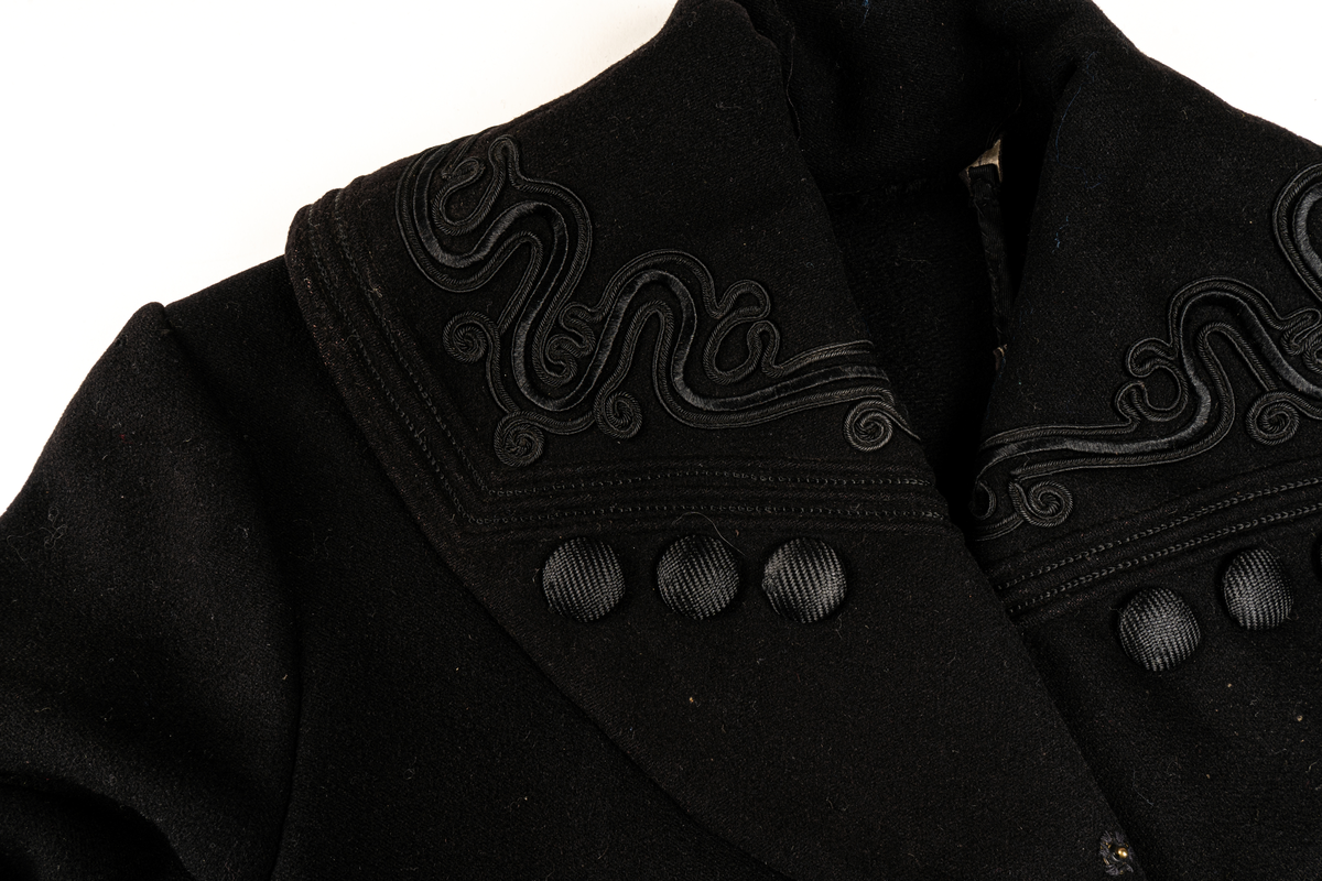 Svart kappa. Sjalkrage med snörbroderier och klädda knappar. Större oklädda knappar framtill.