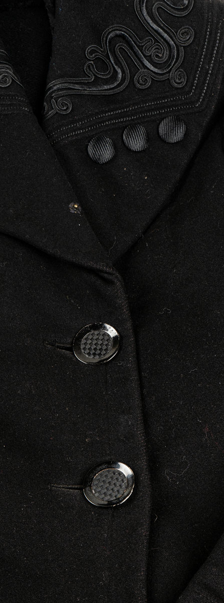 Svart kappa. Sjalkrage med snörbroderier och klädda knappar. Större oklädda knappar framtill.