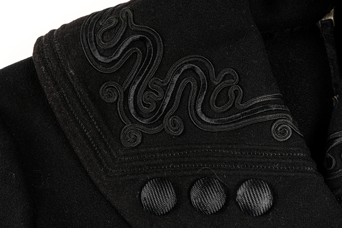 Svart kappa. Sjalkrage med snörbroderier och klädda knappar. Större oklädda knappar framtill.