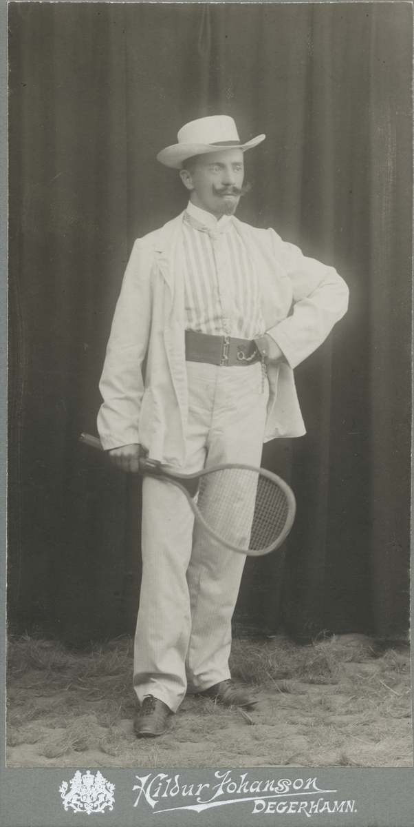Porträtt av man i vita kläder och hatt samt ett tennisracket.