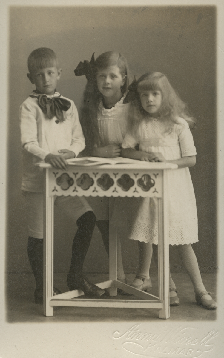 Porträtt av tre barn som står vid ett litet bord.