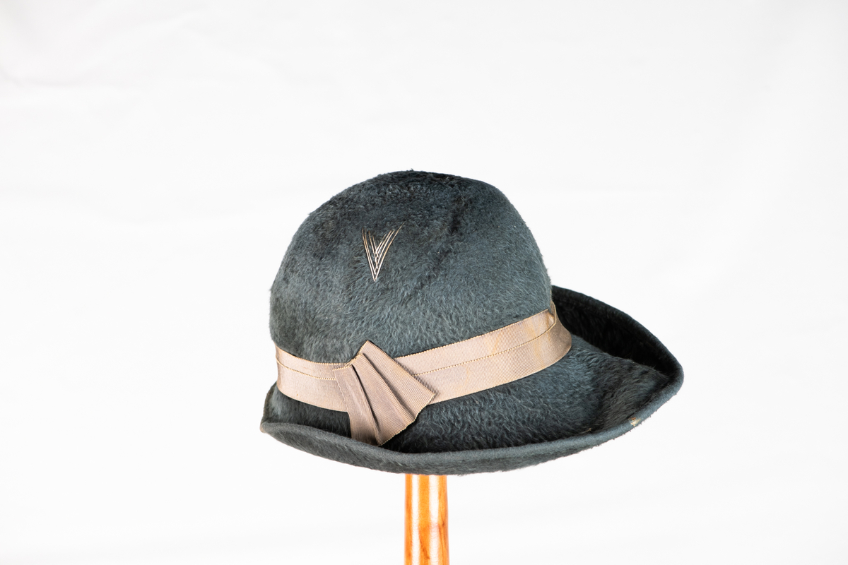 Hatt med brem og pyntebånd (3,3cm.). Bremen er oppbrettet rundt hele hatten, og større i front enn bak. På siden av hatten er det brodert en V-form med tråder.
