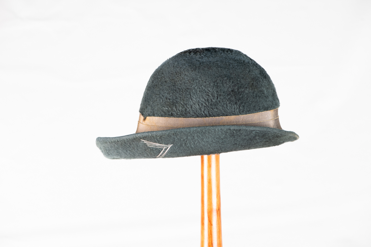 Hatt med brem og pyntebånd (3,3cm.). Bremen er oppbrettet rundt hele hatten, og større i front enn bak. På siden av hatten er det brodert en V-form med tråder.
