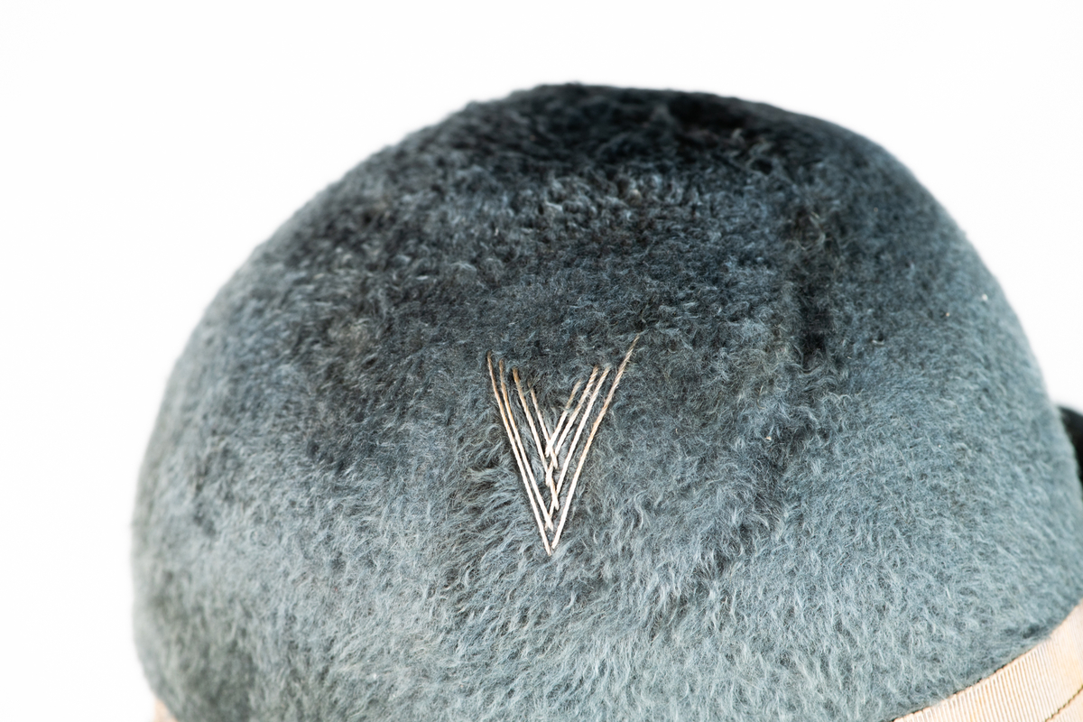 Hatt med brem og pyntebånd (3,3cm.). Bremen er oppbrettet rundt hele hatten, og større i front enn bak. På siden av hatten er det brodert en V-form med tråder.

