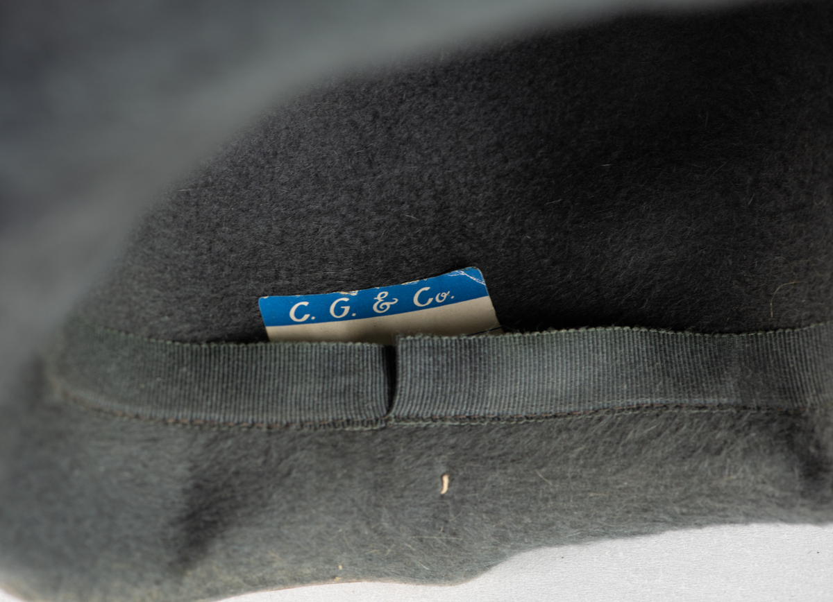 Hatt med brem og pyntebånd (3,3cm.). Bremen er oppbrettet rundt hele hatten, og større i front enn bak. På siden av hatten er det brodert en V-form med tråder.
