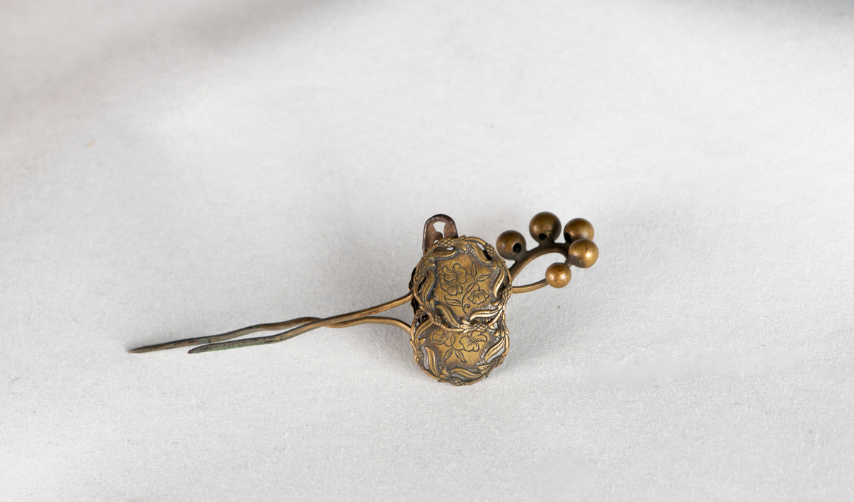 Hattenål i messing. Dekor av fire knapper (mulig fra 1700-tallet)  med blomstergren  med to blomster. Rundt kanten bølgende gren.  Festet til messing bøyle med 7 små kuler på toppen. Gjennom det hele en sikkerhetsnål.