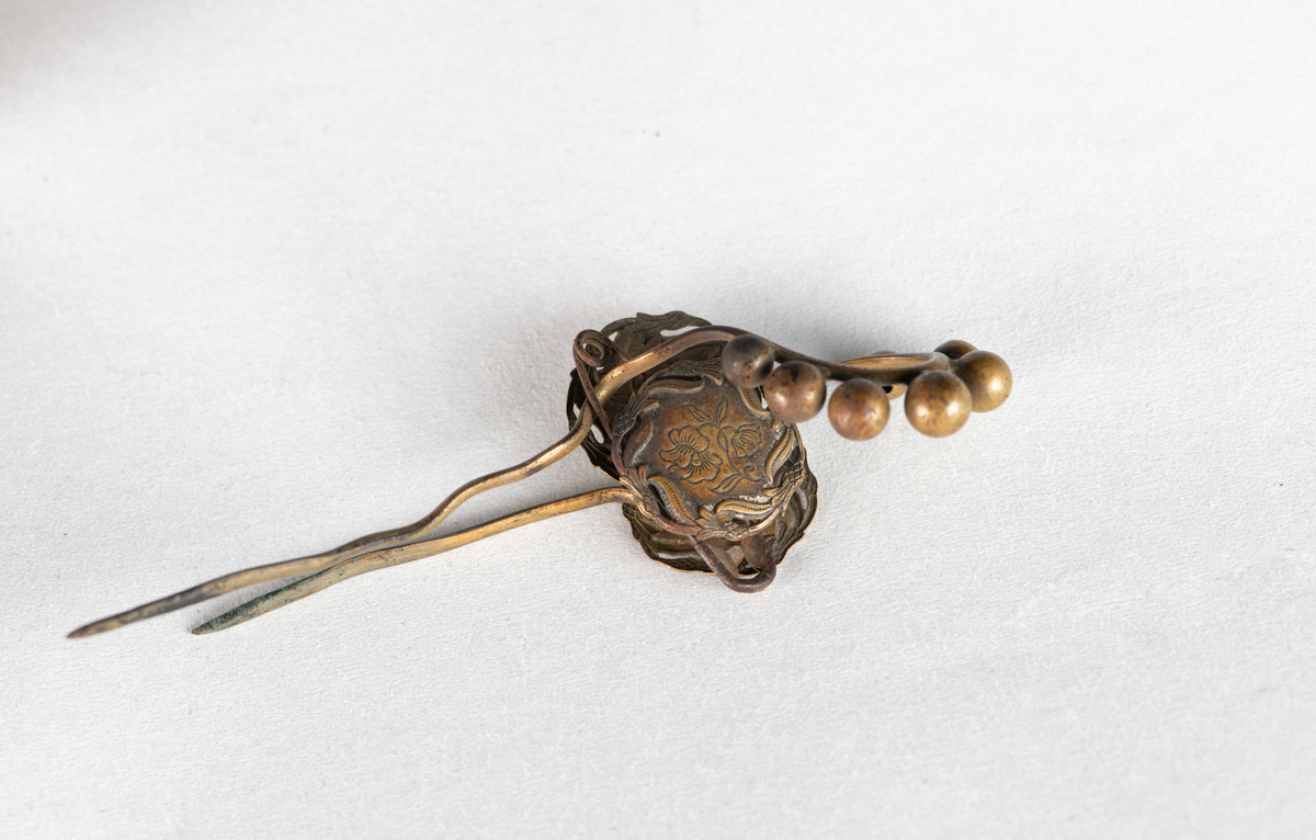 Hattenål i messing. Dekor av fire knapper (mulig fra 1700-tallet)  med blomstergren  med to blomster. Rundt kanten bølgende gren.  Festet til messing bøyle med 7 små kuler på toppen. Gjennom det hele en sikkerhetsnål.