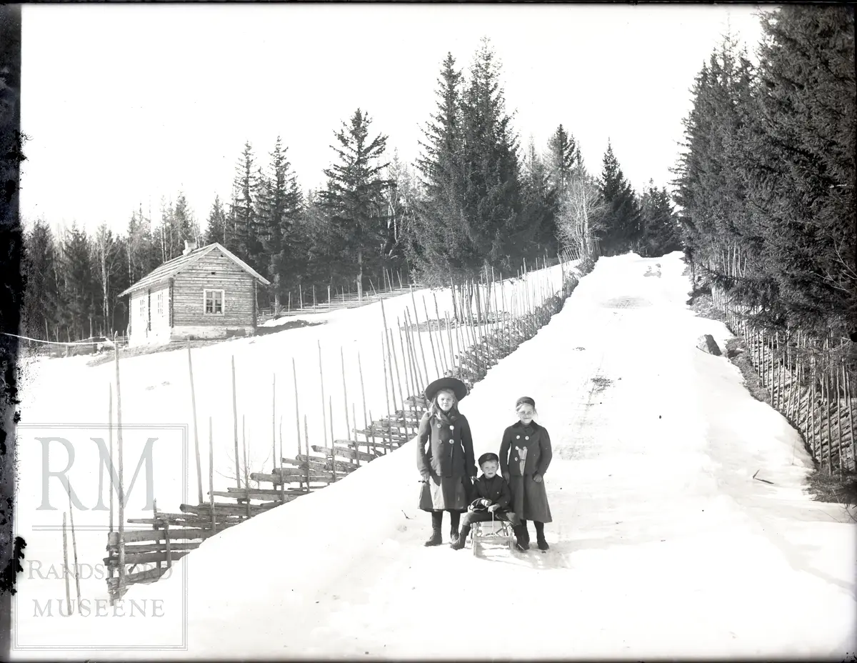 En gutt sitter på en kjelke og to jenter står ved siden av. Snødekket vei med skigard på begge sider. Et hus til venstre i bilde. 