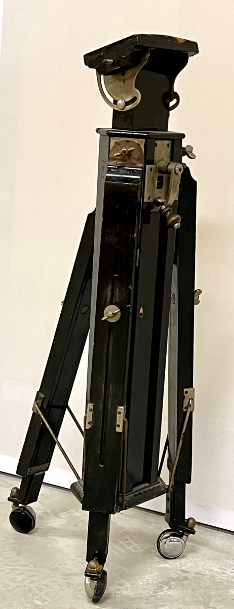 Kraftig stativ i sortlakkert tre med tre føtter som kan reguleres i lengde og vinkel. Det er montert hjul på føttene. Øverst finnes en lite plattform som kan tiltes. Stativet er trolig for montering av et fotografiapparat, lyskilde eller et måleinstrument.