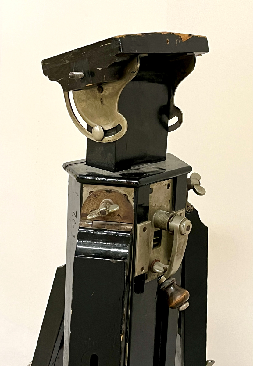 Kraftig stativ i sortlakkert tre med tre føtter som kan reguleres i lengde og vinkel. Det er montert hjul på føttene. Øverst finnes en lite plattform som kan tiltes. Stativet er trolig for montering av et fotografiapparat, lyskilde eller et måleinstrument.