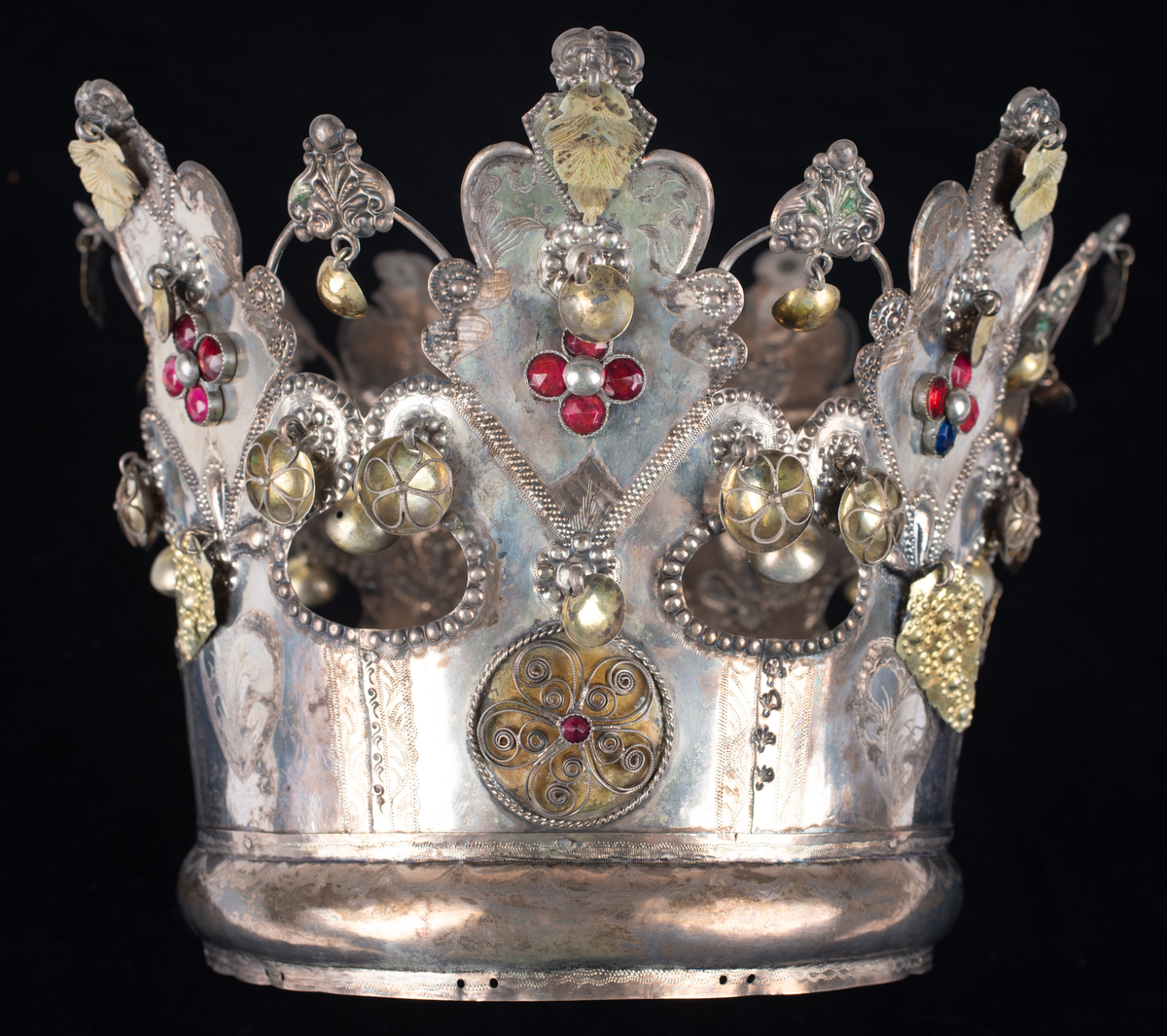 Brudkrona av silverplåt. Oval rak sida av 7 hoplödda plåtar som upptill svänger ut och avslutas i treflikade krön. Buckelornamentik samt svisselerade mönster. Fyrpassformade infattningar av röda och gröna glasstenar på varje flik. Ytterligare dekorerad med påsatta löst hängande förgyllda löv samt halvsfäriska hängen. Kronan avslutas nertill med en påsatt vulst. Stämplar: 13 1/3 (lödighet), LMS (=LM Salvesen), (Ålesunds stadsvapen), 1854.Mått upptill: längd 23 cm, bredd 23 cm.nertill: längd 17 cm, bredd 16 cm, höjd 17 cm.