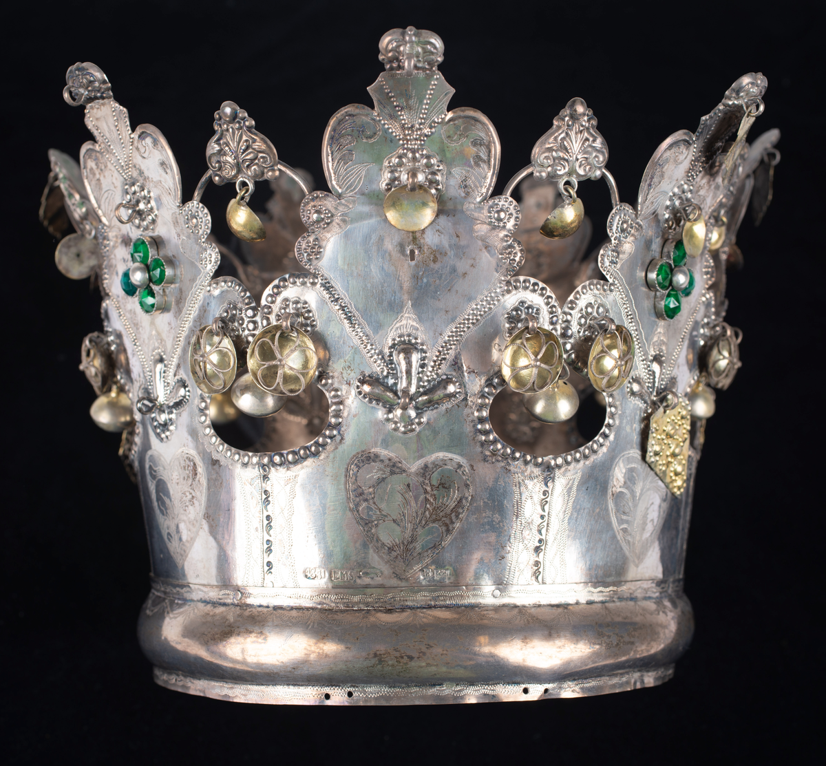 Brudkrona av silverplåt. Oval rak sida av 7 hoplödda plåtar som upptill svänger ut och avslutas i treflikade krön. Buckelornamentik samt svisselerade mönster. Fyrpassformade infattningar av röda och gröna glasstenar på varje flik. Ytterligare dekorerad med påsatta löst hängande förgyllda löv samt halvsfäriska hängen. Kronan avslutas nertill med en påsatt vulst. Stämplar: 13 1/3 (lödighet), LMS (=LM Salvesen), (Ålesunds stadsvapen), 1854.Mått upptill: längd 23 cm, bredd 23 cm.nertill: längd 17 cm, bredd 16 cm, höjd 17 cm.