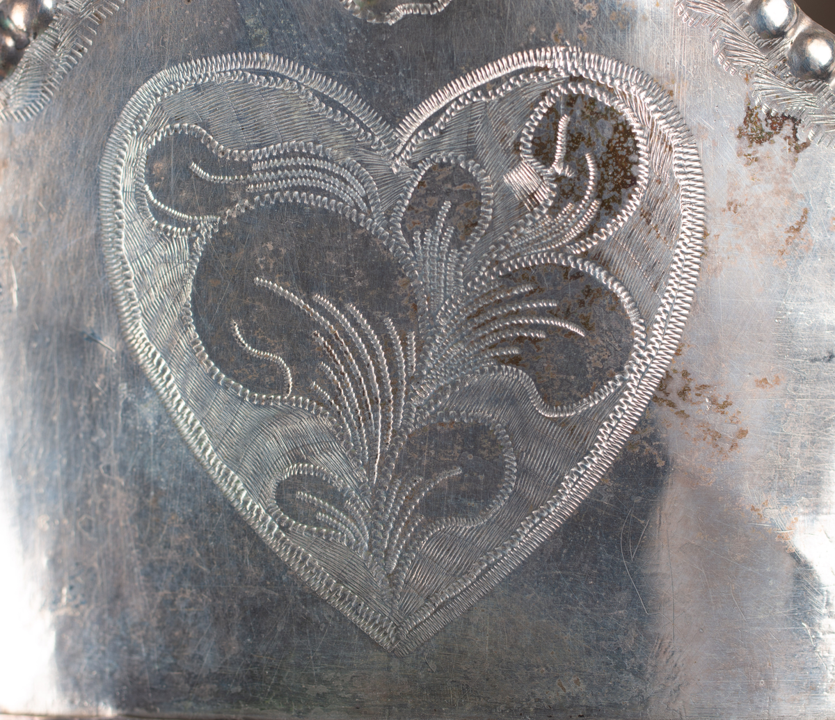 Brudkrona av silverplåt. Oval rak sida av 7 hoplödda plåtar som upptill svänger ut och avslutas i treflikade krön. Buckelornamentik samt svisselerade mönster. Fyrpassformade infattningar av röda och gröna glasstenar på varje flik. Ytterligare dekorerad med påsatta löst hängande förgyllda löv samt halvsfäriska hängen. Kronan avslutas nertill med en påsatt vulst. Stämplar: 13 1/3 (lödighet), LMS (=LM Salvesen), (Ålesunds stadsvapen), 1854.Mått upptill: längd 23 cm, bredd 23 cm.nertill: längd 17 cm, bredd 16 cm, höjd 17 cm.