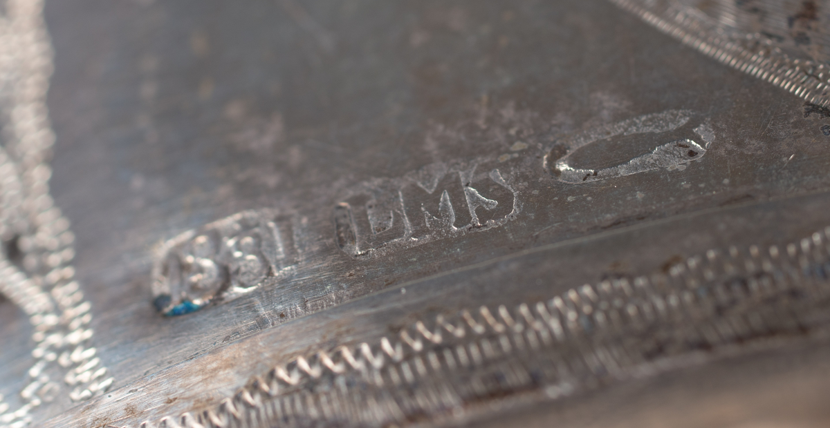 Brudkrona av silverplåt. Oval rak sida av 7 hoplödda plåtar som upptill svänger ut och avslutas i treflikade krön. Buckelornamentik samt svisselerade mönster. Fyrpassformade infattningar av röda och gröna glasstenar på varje flik. Ytterligare dekorerad med påsatta löst hängande förgyllda löv samt halvsfäriska hängen. Kronan avslutas nertill med en påsatt vulst. Stämplar: 13 1/3 (lödighet), LMS (=LM Salvesen), (Ålesunds stadsvapen), 1854.Mått upptill: längd 23 cm, bredd 23 cm.nertill: längd 17 cm, bredd 16 cm, höjd 17 cm.