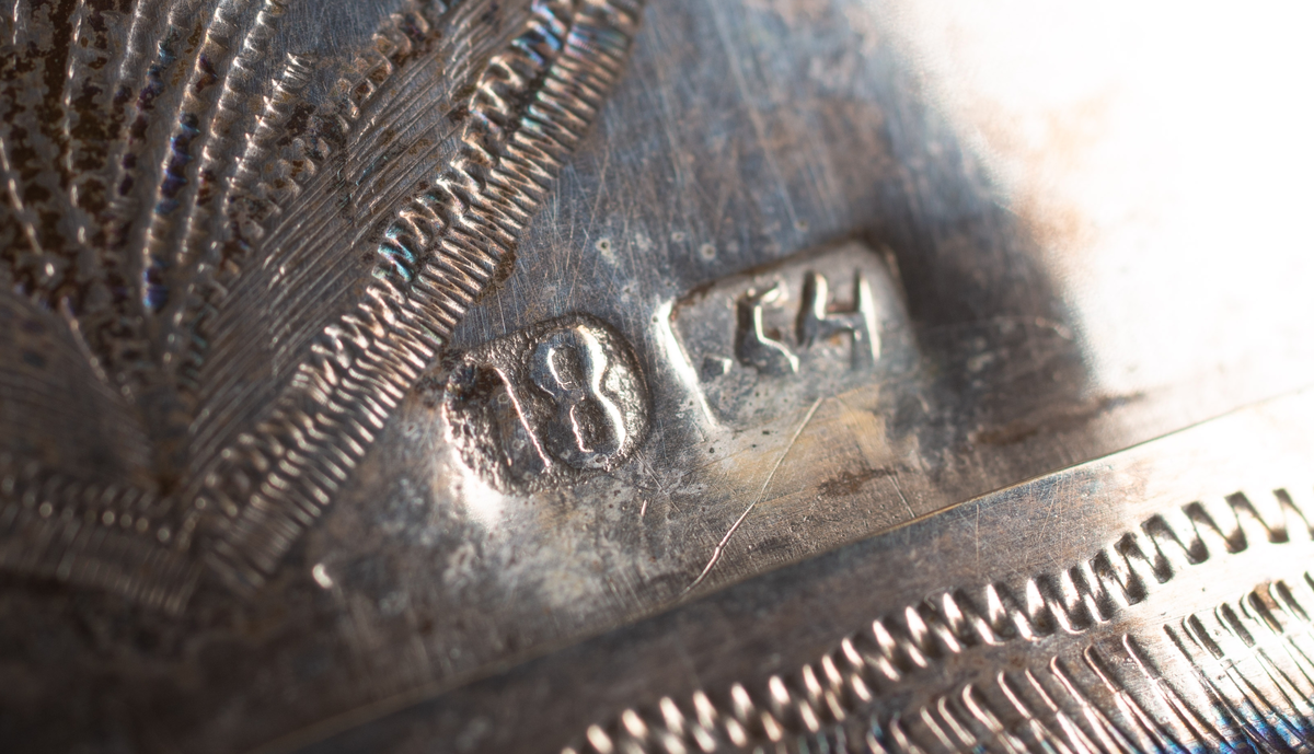 Brudkrona av silverplåt. Oval rak sida av 7 hoplödda plåtar som upptill svänger ut och avslutas i treflikade krön. Buckelornamentik samt svisselerade mönster. Fyrpassformade infattningar av röda och gröna glasstenar på varje flik. Ytterligare dekorerad med påsatta löst hängande förgyllda löv samt halvsfäriska hängen. Kronan avslutas nertill med en påsatt vulst. Stämplar: 13 1/3 (lödighet), LMS (=LM Salvesen), (Ålesunds stadsvapen), 1854.Mått upptill: längd 23 cm, bredd 23 cm.nertill: längd 17 cm, bredd 16 cm, höjd 17 cm.