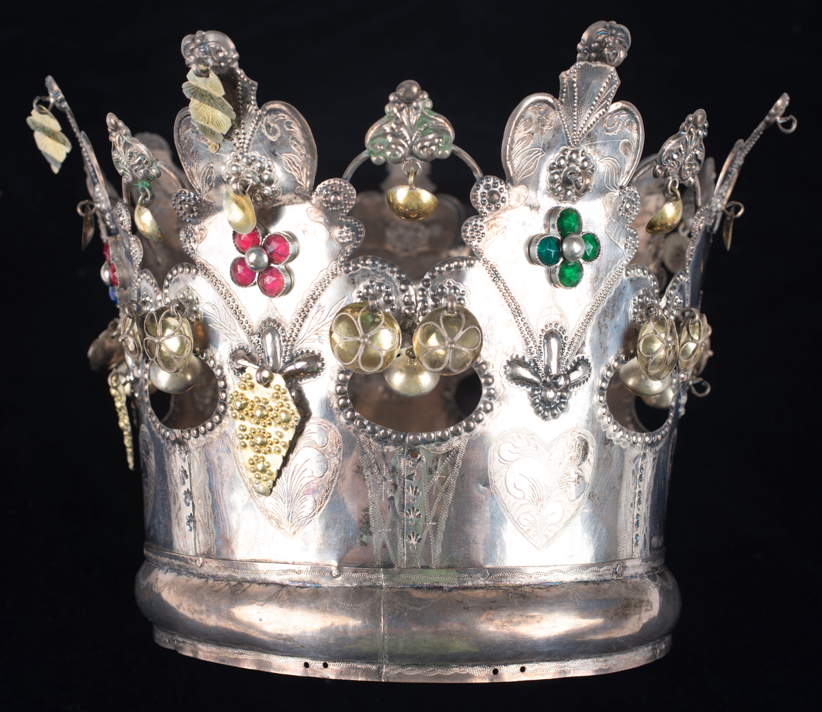 Brudkrona av silverplåt. Oval rak sida av 7 hoplödda plåtar som upptill svänger ut och avslutas i treflikade krön. Buckelornamentik samt svisselerade mönster. Fyrpassformade infattningar av röda och gröna glasstenar på varje flik. Ytterligare dekorerad med påsatta löst hängande förgyllda löv samt halvsfäriska hängen. Kronan avslutas nertill med en påsatt vulst. Stämplar: 13 1/3 (lödighet), LMS (=LM Salvesen), (Ålesunds stadsvapen), 1854.Mått upptill: längd 23 cm, bredd 23 cm.nertill: längd 17 cm, bredd 16 cm, höjd 17 cm.