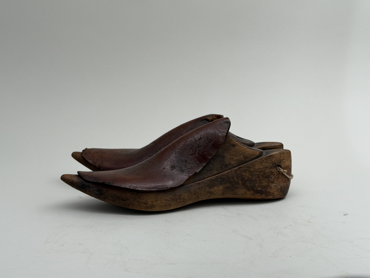 Skoläster, ett par, av björk. Överdelarna fästa med spårskurv, samt överklädda med två bitar brunt läder vardera. Häl med hål för upphängning.