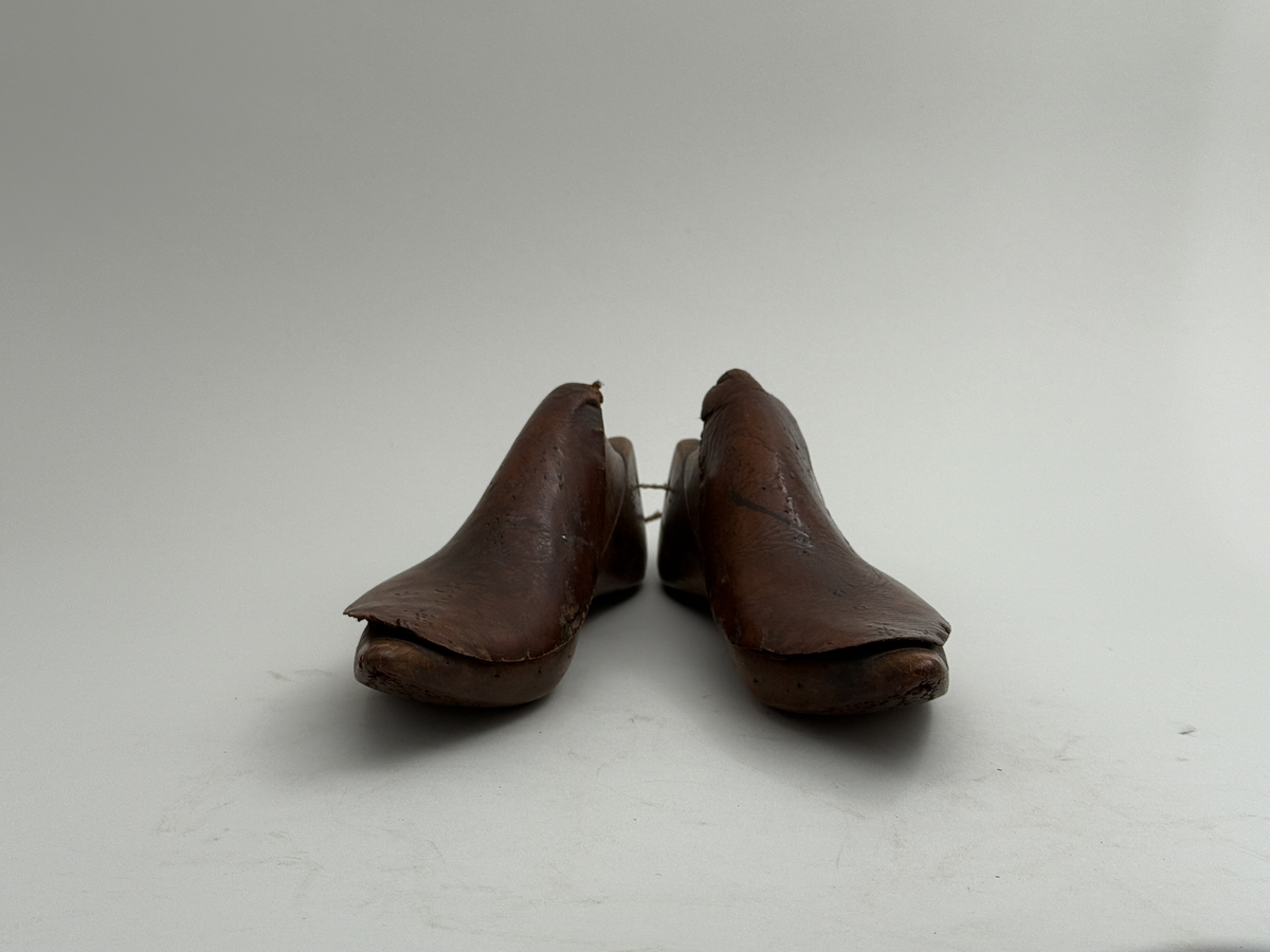 Skoläster, ett par, av björk. Överdelarna fästa med spårskurv, samt överklädda med två bitar brunt läder vardera. Häl med hål för upphängning.