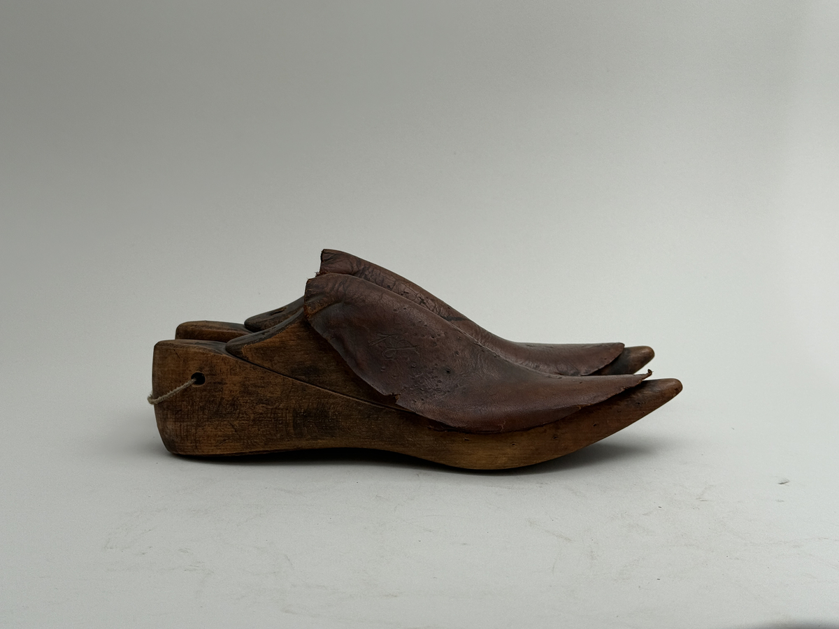 Skoläster, ett par, av björk. Överdelarna fästa med spårskurv, samt överklädda med två bitar brunt läder vardera. Häl med hål för upphängning.