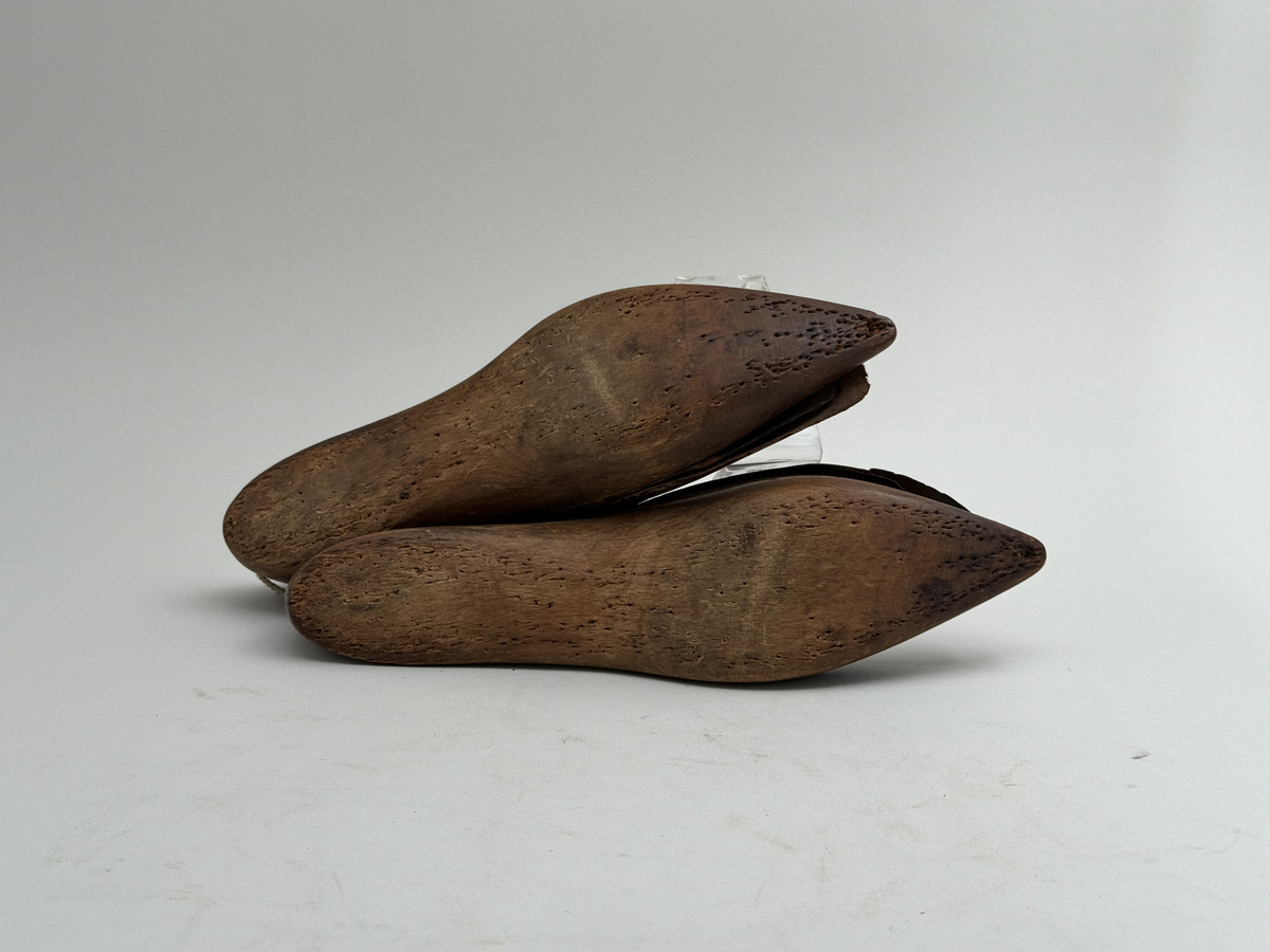 Skoläster, ett par, av björk. Överdelarna fästa med spårskurv, samt överklädda med två bitar brunt läder vardera. Häl med hål för upphängning.