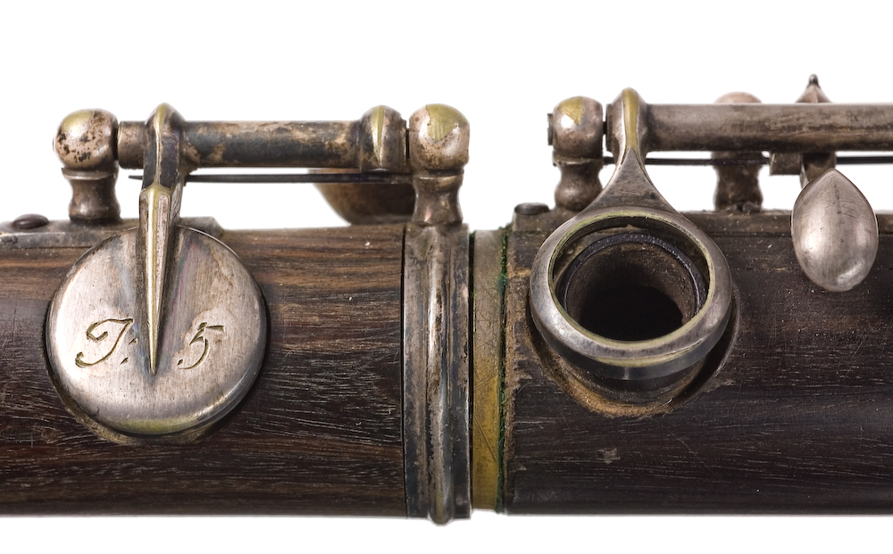 Tvärflöjt i mörkt trä med metallringar. Böhmsystem, delvis med öppna klaffar. 3 delar. Sprickor i munstycke. Fot ej original. Dålig kondition. A=459.