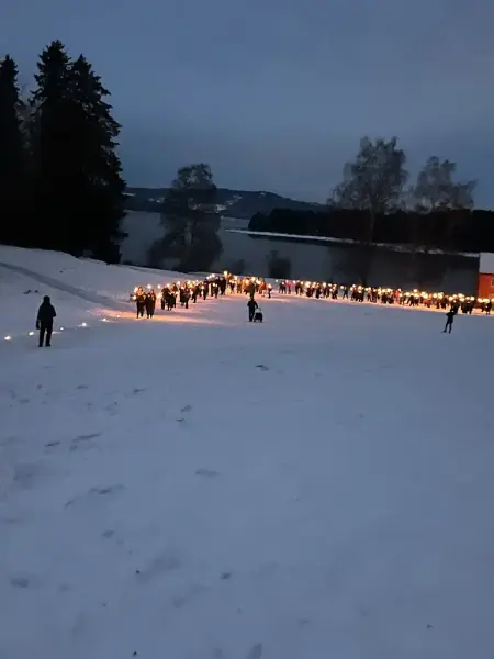I et snødekket landskap går en lang rekke mennesker med tente fakler på Domkirkeodden, Mjøsa i bakgrunnen.