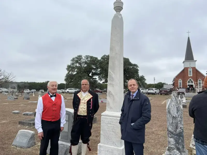 Tre menn står ved et høyt gravmonument. De to til venstre har på bunad. En kirke i bakgrunn til høyre. 