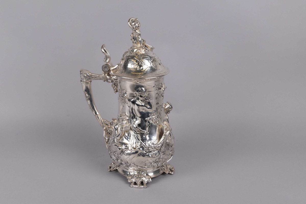 Kanne dekorert med gjennomgådene relieffmønster av figurer og kjeruber blant vinranker og druer. Kuppellokk med motiv av en kjerub med banjo som skåler med et glass vin. Håndtaket dekorert med vinranker og druer.