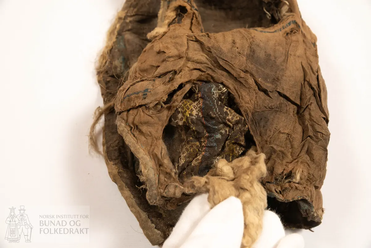 Lue, tostykkslue. Sauma av mørk brun lerret i bomullskvalitet (?), med flotterande trådar som dannar blomemønster, i innslagsretning. Vekslande fargeparti i blått, lysblått, rosa, raudt, gult og grønt - taboret. For av tynn silkelerret, stripevove. Mellomfor av litt tjukk bomullsvatt, stukke til silkeforet med ca 4-5 cm stingavstand. Kanting langs framkant av truleg lysblått band av silketaft. Nedkant falda inn begge stoff, og sydd til med faldesting frå innsida. Samansying av hovudstoffa med 1 cm saummon. Venstre fòrdel er falda inn over høgre, og sydd ned med faldesting med kvit tynnare lintråd. Hol etter snøring langs heile nedkant, hola har sårkant gjennom begge lag. I nedkant mot kvar framkant er påsydd ein firkant av grovare bomullsvare, truleg reparasjon.