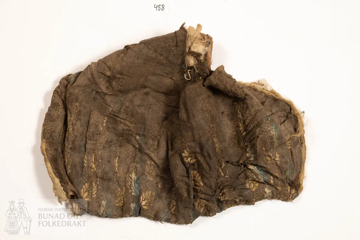 Lue, tostykkslue. Sauma av mørk brun lerret i bomullskvalitet (?), med flotterande trådar som dannar blomemønster, i innslagsretning. Vekslande fargeparti i blått, lysblått, rosa, raudt, gult og grønt - taboret. For av tynn silkelerret, stripevove. Mellomfor av litt tjukk bomullsvatt, stukke til silkeforet med ca 4-5 cm stingavstand. Kanting langs framkant av truleg lysblått band av silketaft. Nedkant falda inn begge stoff, og sydd til med faldesting frå innsida. Samansying av hovudstoffa med 1 cm saummon. Venstre fòrdel er falda inn over høgre, og sydd ned med faldesting med kvit tynnare lintråd. Hol etter snøring langs heile nedkant, hola har sårkant gjennom begge lag. I nedkant mot kvar framkant er påsydd ein firkant av grovare bomullsvare, truleg reparasjon.