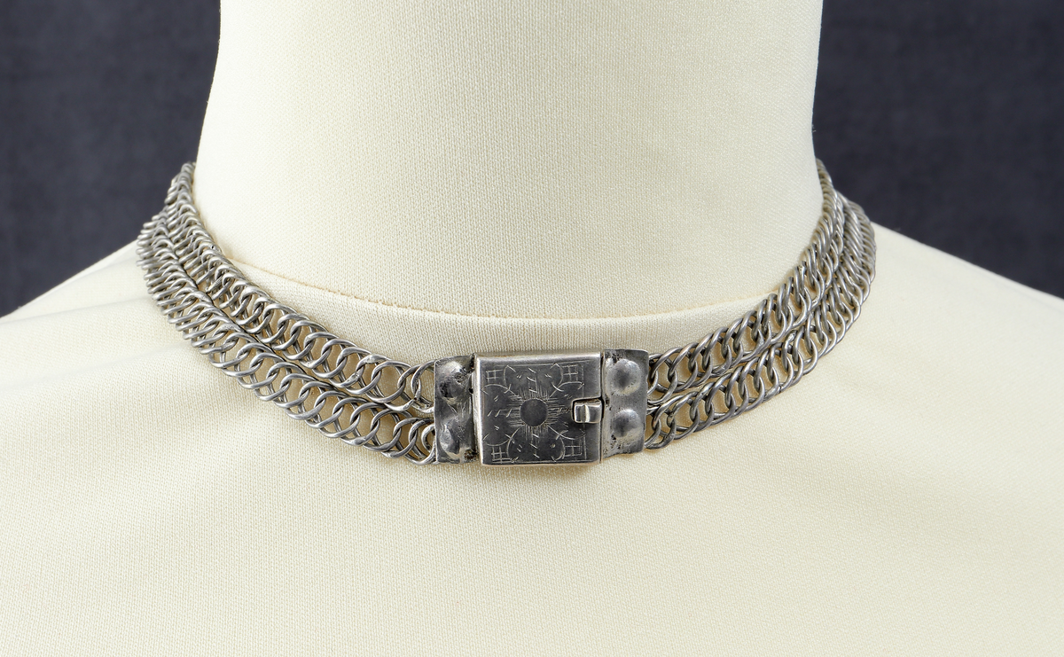 Halslås av silver. Ostämplat. Smycket är tillverkat  som en grund silverdosa med därpå fästade kedjor av pansartyp. Det låses genom en dubbelvikt fjädrande silverplåt som skjuts in från sida.- Dosan har gravyr, föreställande en stiliserad blomma på rutmönster.

Inskrivet i huvudkatalog 1938.