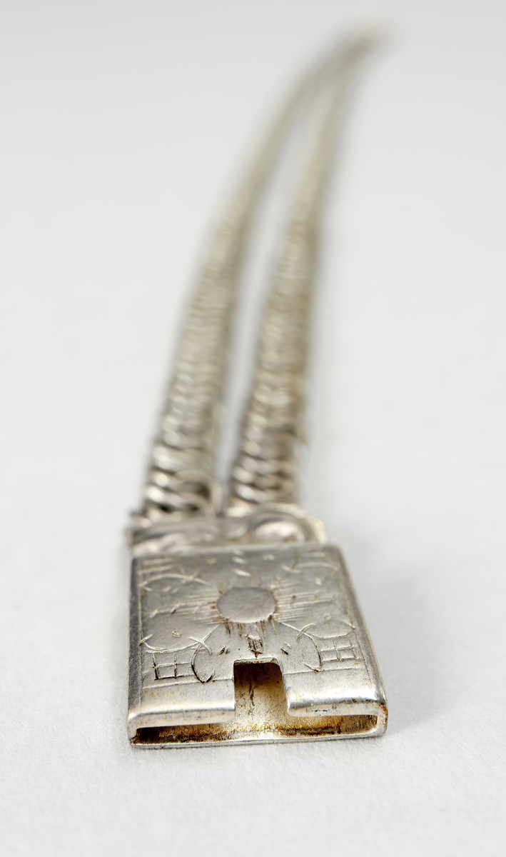 Halslås av silver. Ostämplat. Smycket är tillverkat  som en grund silverdosa med därpå fästade kedjor av pansartyp. Det låses genom en dubbelvikt fjädrande silverplåt som skjuts in från sida.- Dosan har gravyr, föreställande en stiliserad blomma på rutmönster.

Inskrivet i huvudkatalog 1938.