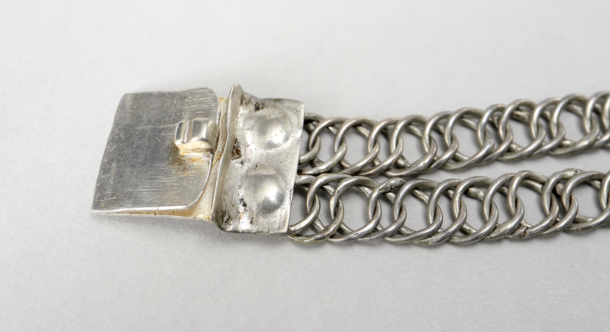 Halslås av silver. Ostämplat. Smycket är tillverkat  som en grund silverdosa med därpå fästade kedjor av pansartyp. Det låses genom en dubbelvikt fjädrande silverplåt som skjuts in från sida.- Dosan har gravyr, föreställande en stiliserad blomma på rutmönster.

Inskrivet i huvudkatalog 1938.