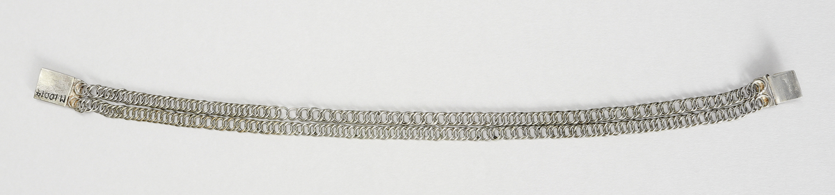 Halslås av silver. Ostämplat. Smycket är tillverkat  som en grund silverdosa med därpå fästade kedjor av pansartyp. Det låses genom en dubbelvikt fjädrande silverplåt som skjuts in från sida.- Dosan har gravyr, föreställande en stiliserad blomma på rutmönster.

Inskrivet i huvudkatalog 1938.