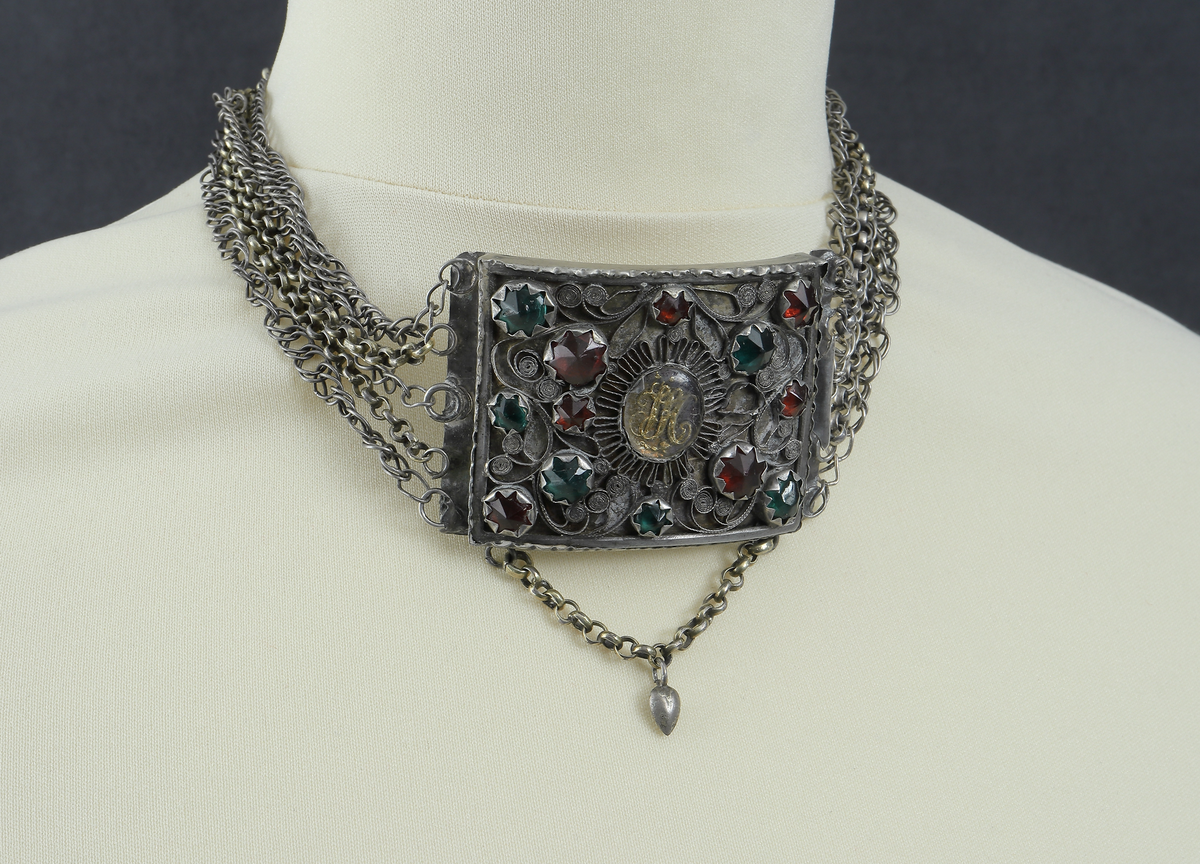 Halslås av silver. Består av en välvd fyrkantig dosa. På framsidan är dekoren gjord i filigranarbete samt besatt med gröna och röda fasseterade glasflusser. I mitten finns ett namnchiffer under en oval infattad glasbit. Chiffret består av bokstäverna J.H.S. (Jesus Hominum Salvator). Plattan under filigranet har varit förgyllt. I halslåsets ytterkanter sitter fem kedjor infästa. På den ena sidan sitter låsanordningen. Kedjorna är utförda av pansar- och ankartyp. I halslåsets underkant hänger en liten kedja med ett spetsigt silverhjärta.
Inskrivet i huvudkatalog 1950.