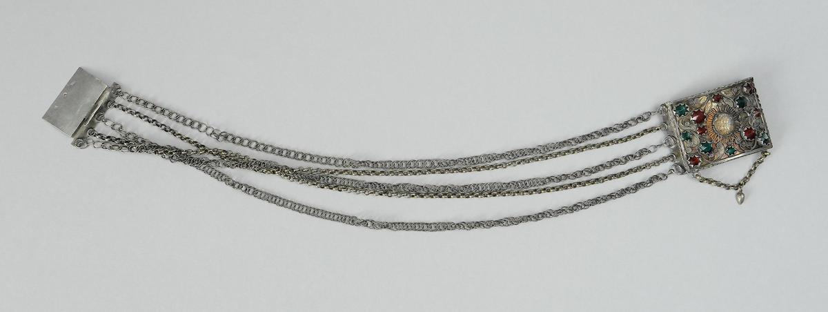 Halslås av silver. Består av en välvd fyrkantig dosa. På framsidan är dekoren gjord i filigranarbete samt besatt med gröna och röda fasseterade glasflusser. I mitten finns ett namnchiffer under en oval infattad glasbit. Chiffret består av bokstäverna J.H.S. (Jesus Hominum Salvator). Plattan under filigranet har varit förgyllt. I halslåsets ytterkanter sitter fem kedjor infästa. På den ena sidan sitter låsanordningen. Kedjorna är utförda av pansar- och ankartyp. I halslåsets underkant hänger en liten kedja med ett spetsigt silverhjärta.
Inskrivet i huvudkatalog 1950.