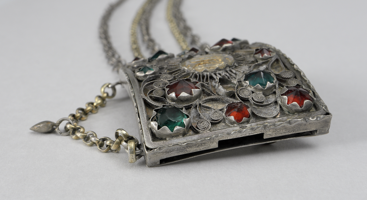 Halslås av silver. Består av en välvd fyrkantig dosa. På framsidan är dekoren gjord i filigranarbete samt besatt med gröna och röda fasseterade glasflusser. I mitten finns ett namnchiffer under en oval infattad glasbit. Chiffret består av bokstäverna J.H.S. (Jesus Hominum Salvator). Plattan under filigranet har varit förgyllt. I halslåsets ytterkanter sitter fem kedjor infästa. På den ena sidan sitter låsanordningen. Kedjorna är utförda av pansar- och ankartyp. I halslåsets underkant hänger en liten kedja med ett spetsigt silverhjärta.
Inskrivet i huvudkatalog 1950.