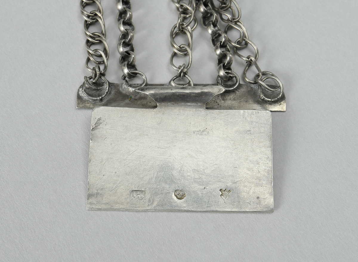 Halslås av silver. Består av en välvd fyrkantig dosa. På framsidan är dekoren gjord i filigranarbete samt besatt med gröna och röda fasseterade glasflusser. I mitten finns ett namnchiffer under en oval infattad glasbit. Chiffret består av bokstäverna J.H.S. (Jesus Hominum Salvator). Plattan under filigranet har varit förgyllt. I halslåsets ytterkanter sitter fem kedjor infästa. På den ena sidan sitter låsanordningen. Kedjorna är utförda av pansar- och ankartyp. I halslåsets underkant hänger en liten kedja med ett spetsigt silverhjärta.
Inskrivet i huvudkatalog 1950.