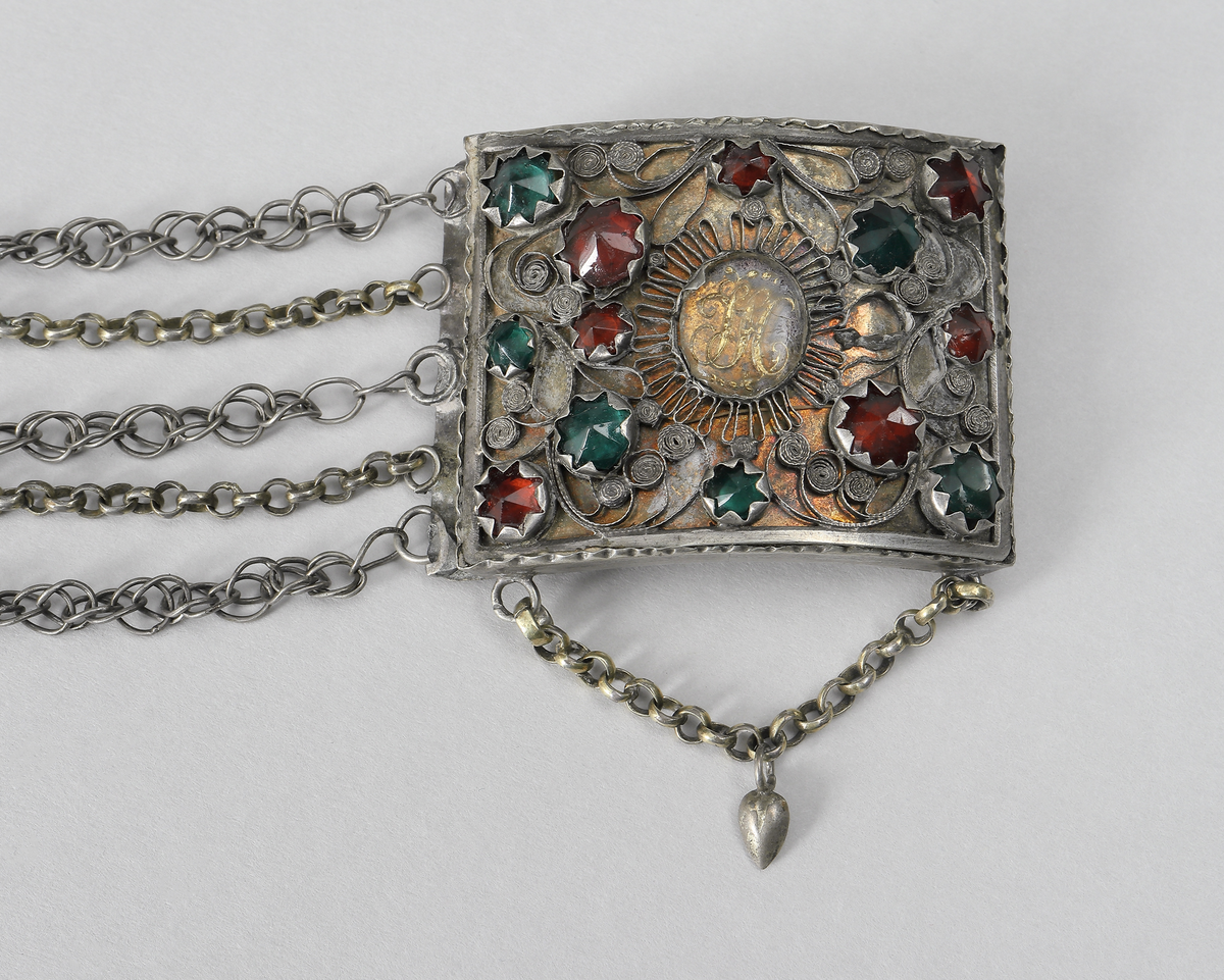 Halslås av silver. Består av en välvd fyrkantig dosa. På framsidan är dekoren gjord i filigranarbete samt besatt med gröna och röda fasseterade glasflusser. I mitten finns ett namnchiffer under en oval infattad glasbit. Chiffret består av bokstäverna J.H.S. (Jesus Hominum Salvator). Plattan under filigranet har varit förgyllt. I halslåsets ytterkanter sitter fem kedjor infästa. På den ena sidan sitter låsanordningen. Kedjorna är utförda av pansar- och ankartyp. I halslåsets underkant hänger en liten kedja med ett spetsigt silverhjärta.
Inskrivet i huvudkatalog 1950.