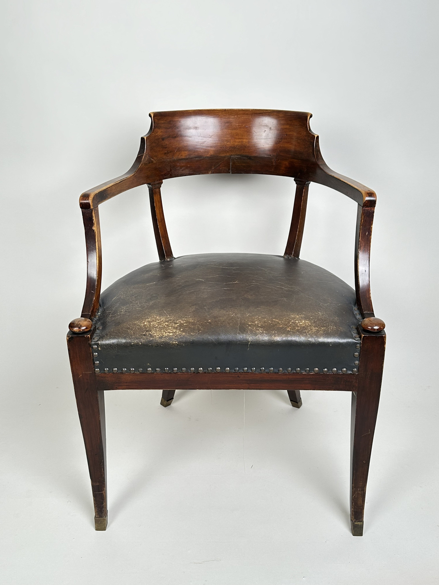 Stol, karmstol. Av polerad, mörkt betsad björk. Mjuk, rundad form med slät ryggbricka, så när som på små skurna volut i dess övre hörn. Ryggståndarna dekorerade med kapitäl i mötet med ryggbrickan. Främre stolsbenen med kulavslut upptill. Samtliga stolsben aningen svängda samt skodda med mässingskor. Sits stoppad och klädd med mörkt grå (troligtvis) pergamoid, fäst med runda, släta nitar. Sekundär sadelgjord undertill av fastnålade tygstycken.