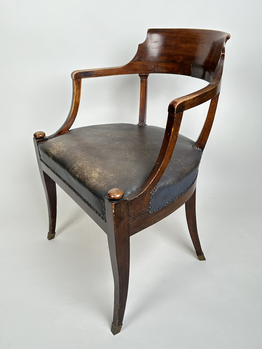 Stol, karmstol. Av polerad, mörkt betsad björk. Mjuk, rundad form med slät ryggbricka, så när som på små skurna volut i dess övre hörn. Ryggståndarna dekorerade med kapitäl i mötet med ryggbrickan. Främre stolsbenen med kulavslut upptill. Samtliga stolsben aningen svängda samt skodda med mässingskor. Sits stoppad och klädd med mörkt grå (troligtvis) pergamoid, fäst med runda, släta nitar. Sekundär sadelgjord undertill av fastnålade tygstycken.