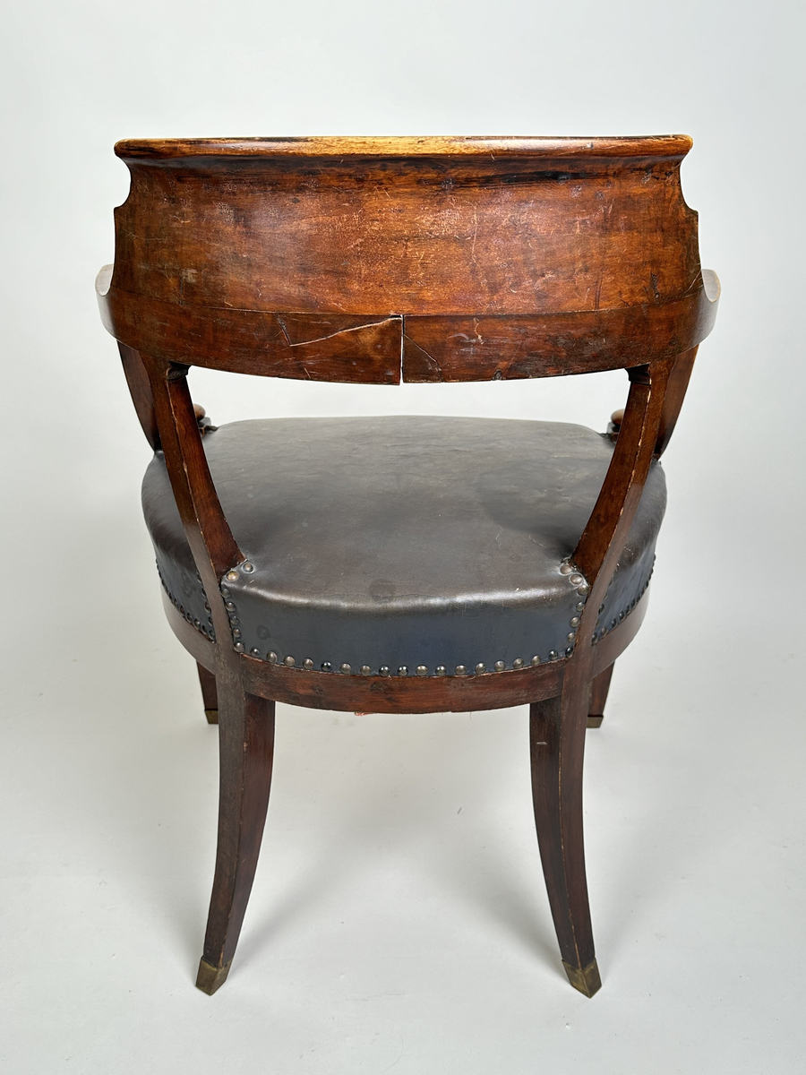 Stol, karmstol. Av polerad, mörkt betsad björk. Mjuk, rundad form med slät ryggbricka, så när som på små skurna volut i dess övre hörn. Ryggståndarna dekorerade med kapitäl i mötet med ryggbrickan. Främre stolsbenen med kulavslut upptill. Samtliga stolsben aningen svängda samt skodda med mässingskor. Sits stoppad och klädd med mörkt grå (troligtvis) pergamoid, fäst med runda, släta nitar. Sekundär sadelgjord undertill av fastnålade tygstycken.