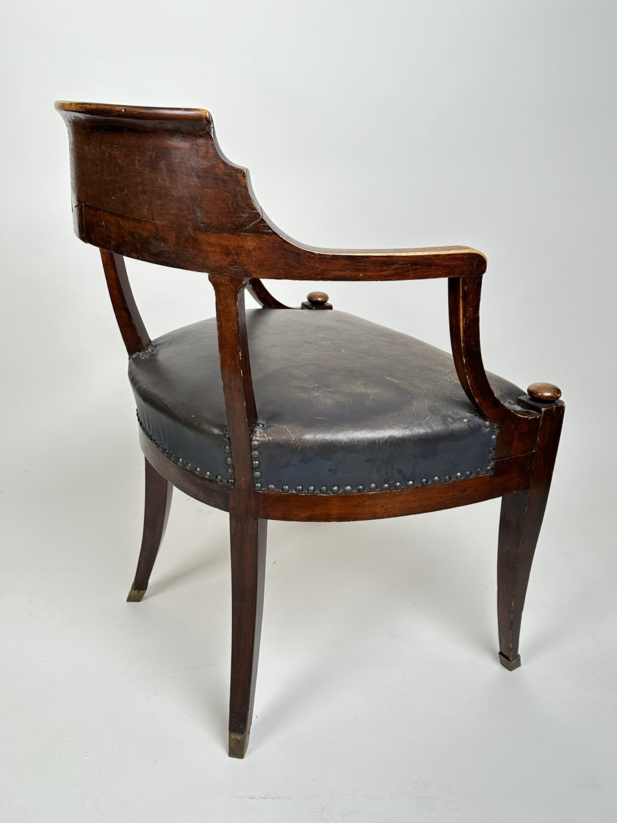 Stol, karmstol. Av polerad, mörkt betsad björk. Mjuk, rundad form med slät ryggbricka, så när som på små skurna volut i dess övre hörn. Ryggståndarna dekorerade med kapitäl i mötet med ryggbrickan. Främre stolsbenen med kulavslut upptill. Samtliga stolsben aningen svängda samt skodda med mässingskor. Sits stoppad och klädd med mörkt grå (troligtvis) pergamoid, fäst med runda, släta nitar. Sekundär sadelgjord undertill av fastnålade tygstycken.