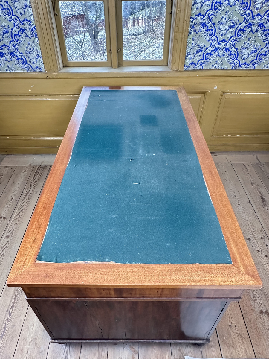 Bordsskiva med sarg till skrivbord, av fernissat och polerat lövträ samt blindträ av furu. Rektangulärt blad klätt med grön filt. Underliggande sarg med plats för extraskiva, samt fem utdragbara lådor - tre på skrivarsidan och två på andra. Samtliga lådor med handtag i mässing samt nyckelskylt formad som en sköld, av vitt material - möjligtvis elfenben.