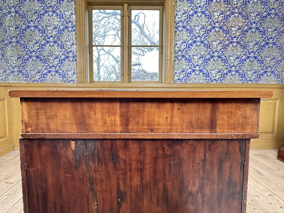 Bordsskiva med sarg till skrivbord, av fernissat och polerat lövträ samt blindträ av furu. Rektangulärt blad klätt med grön filt. Underliggande sarg med plats för extraskiva, samt fem utdragbara lådor - tre på skrivarsidan och två på andra. Samtliga lådor med handtag i mässing samt nyckelskylt formad som en sköld, av vitt material - möjligtvis elfenben.