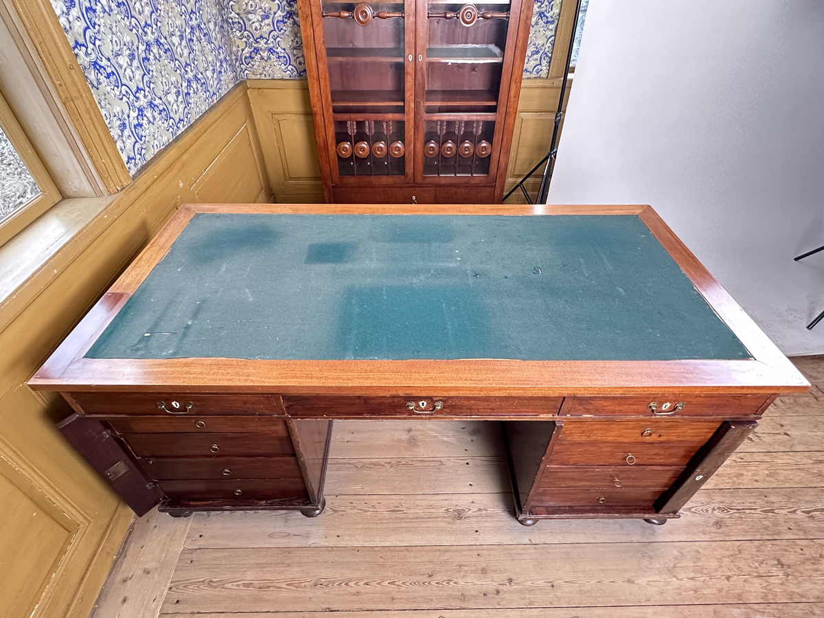 Bordsskiva med sarg till skrivbord, av fernissat och polerat lövträ samt blindträ av furu. Rektangulärt blad klätt med grön filt. Underliggande sarg med plats för extraskiva, samt fem utdragbara lådor - tre på skrivarsidan och två på andra. Samtliga lådor med handtag i mässing samt nyckelskylt formad som en sköld, av vitt material - möjligtvis elfenben.