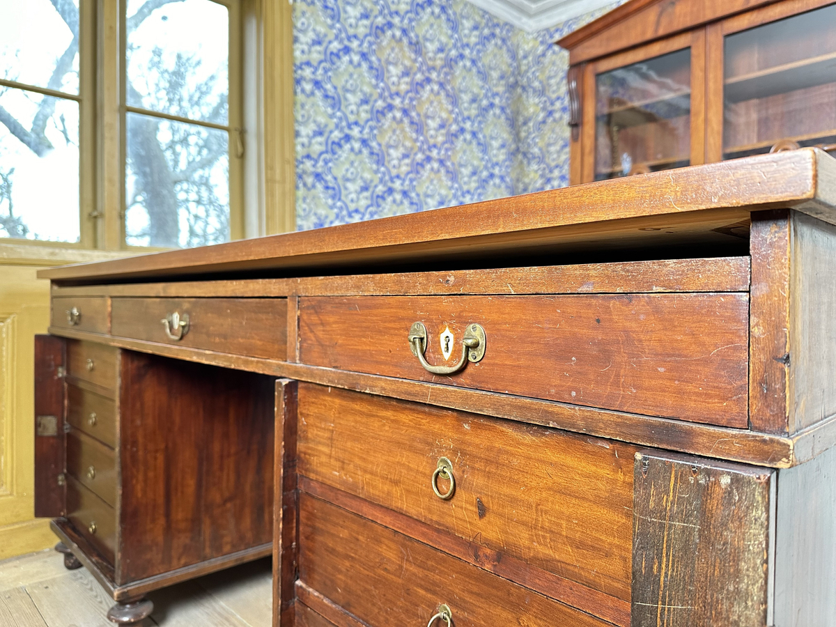 Bordsskiva med sarg till skrivbord, av fernissat och polerat lövträ samt blindträ av furu. Rektangulärt blad klätt med grön filt. Underliggande sarg med plats för extraskiva, samt fem utdragbara lådor - tre på skrivarsidan och två på andra. Samtliga lådor med handtag i mässing samt nyckelskylt formad som en sköld, av vitt material - möjligtvis elfenben.