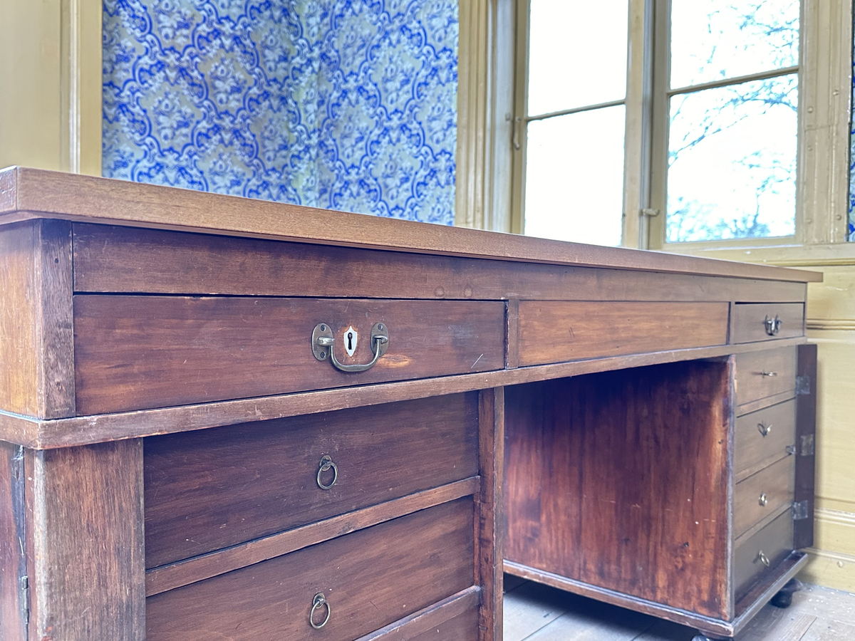 Bordsskiva med sarg till skrivbord, av fernissat och polerat lövträ samt blindträ av furu. Rektangulärt blad klätt med grön filt. Underliggande sarg med plats för extraskiva, samt fem utdragbara lådor - tre på skrivarsidan och två på andra. Samtliga lådor med handtag i mässing samt nyckelskylt formad som en sköld, av vitt material - möjligtvis elfenben.