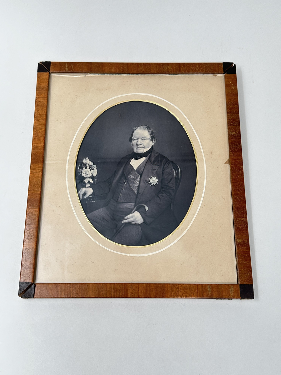 Fotografi i fyrkantig ram av trä med svarta hörn. Fotografiet föreställandes en sittande man i 70-årsåldern, uppklädd med ett ordenstecken på kavajen och käpp i handen. Naturfärgad passepartout med oval öppning för fotografiet, rand i guld och vitt. Baksidan i papp. 
Ramen trasig i hörnen, motivet ligger löst. Glaset i ramen ojämnt klippt upptill. 